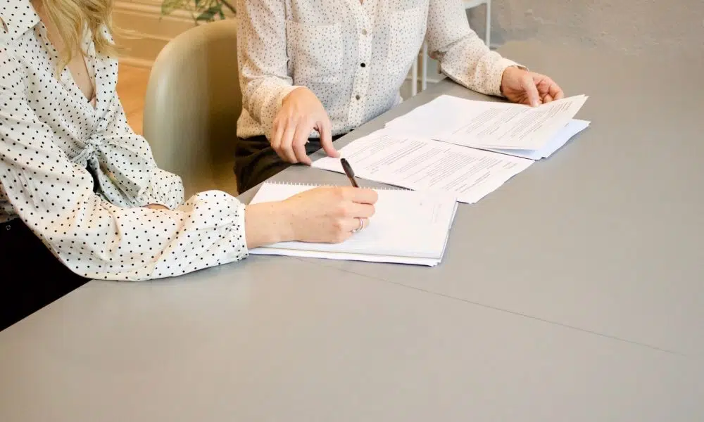 woman signing on white printer paper beside woman about to touch the documents