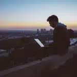 man sitting on concrete brick with opened laptop on his lap